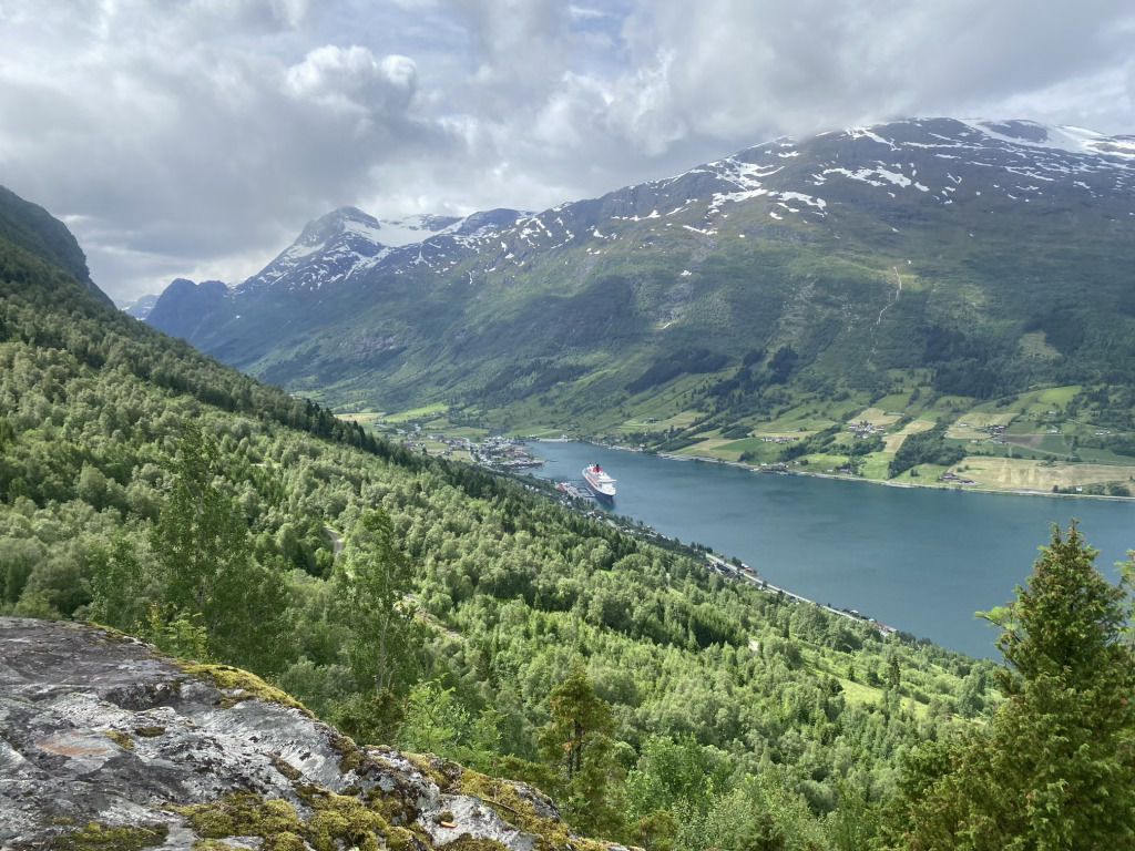 Luftfoto av Huaren-turstien over Olden, Norge, med den skogkledde åssiden, Nordfjorden og snødekte fjell i bakgrunnen