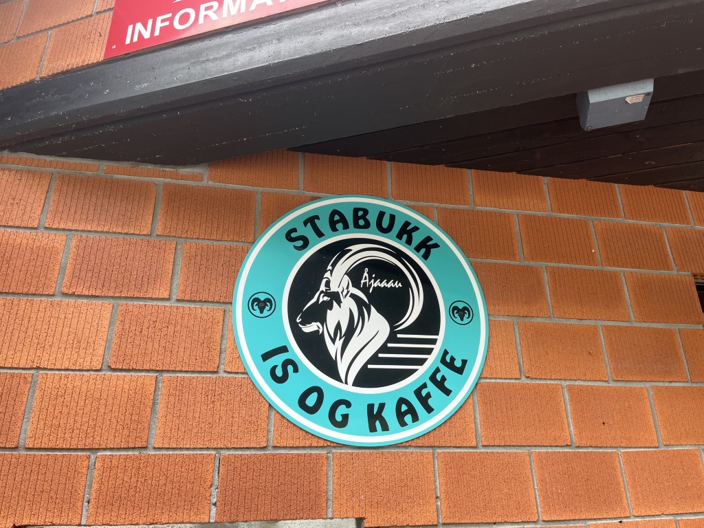 Det turkise Stabukk-skiltet utenfor kafeen i Olden. Midten av skiltet inneholder en vær med store horn. Skiltet er montert på en tradisjonell rød murvegg.