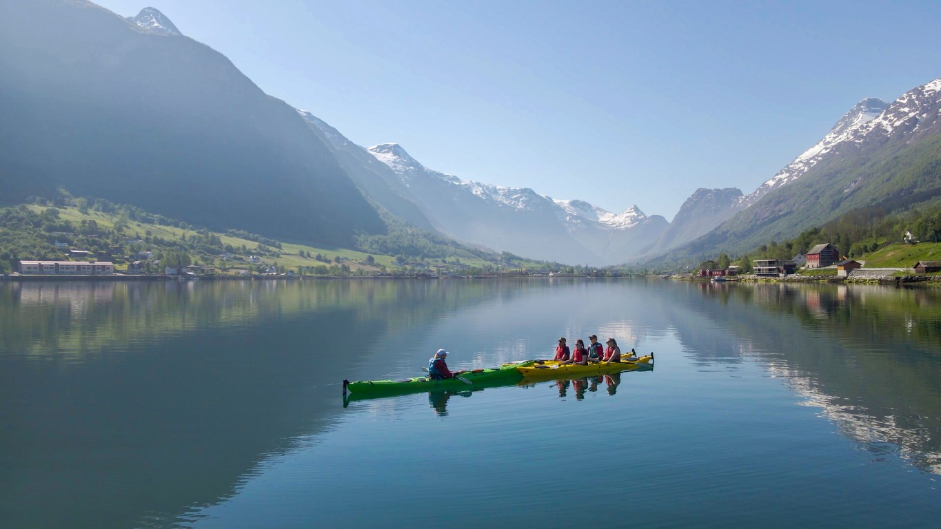 Syklister sykler langs den naturskjønne veien langs innsjøen Oldevatnet i Olden, Norge, med snødekte fjell og turkis vann