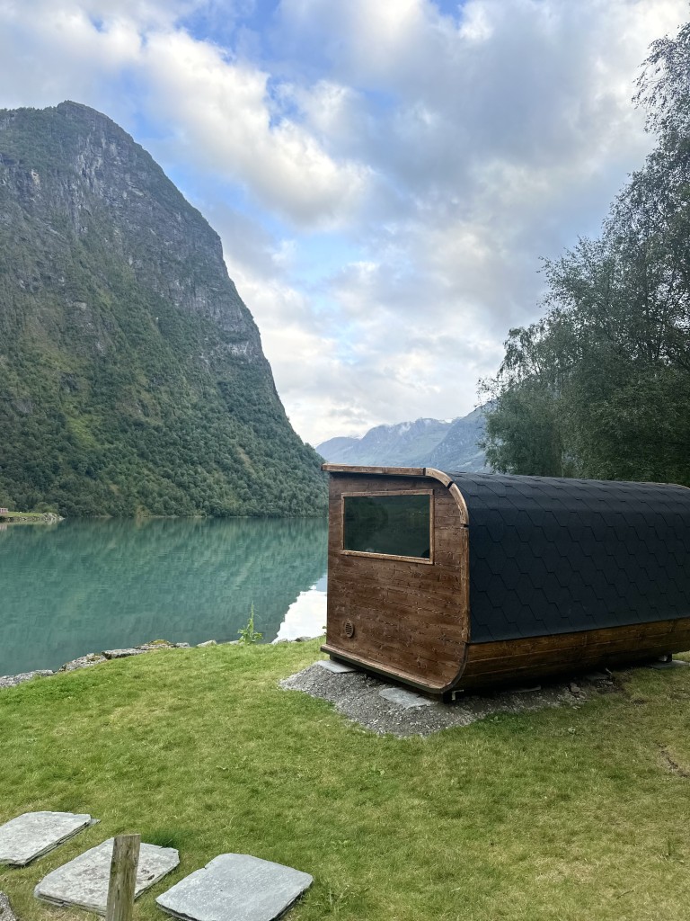 Sidevisning av utendørs badstuhytte i tre ved bredden av Oldevatnet i Olden, Norge, omgitt av dramatiske fjordfjell