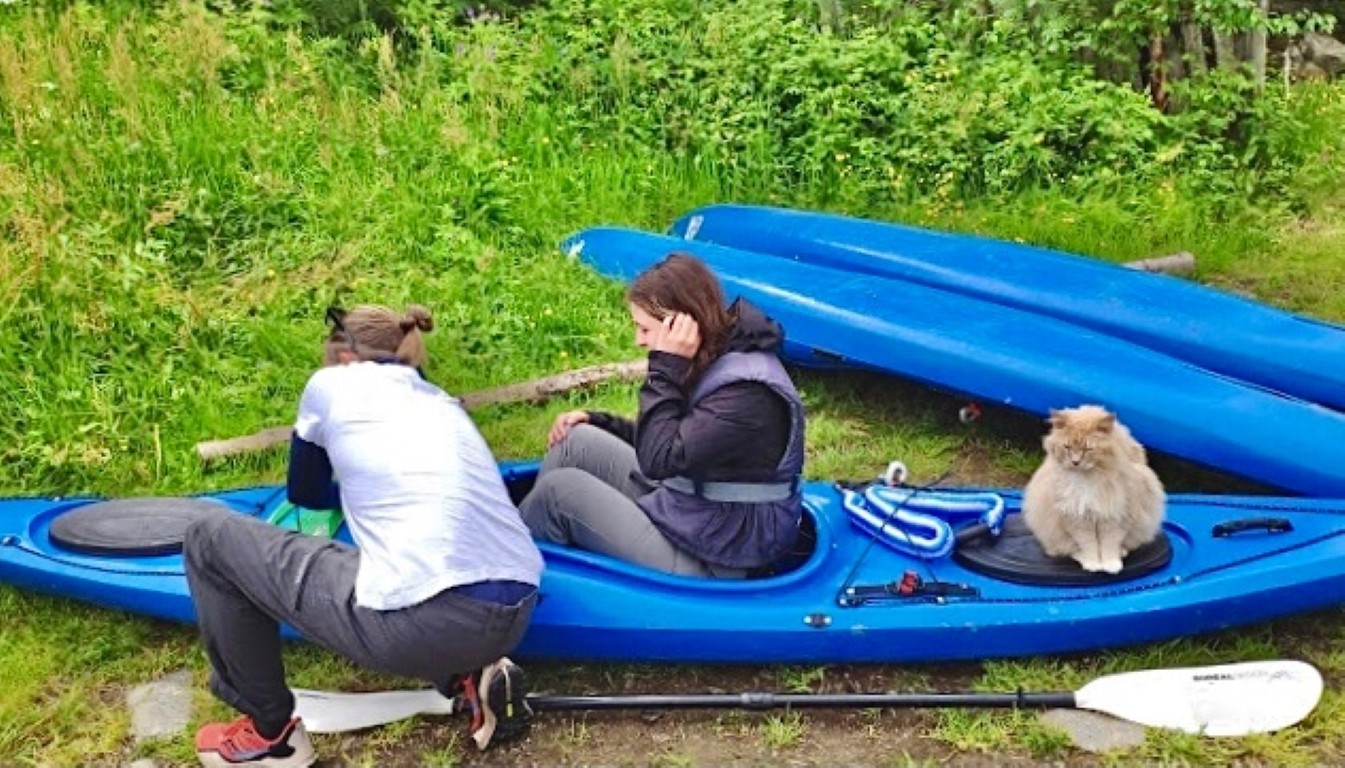 Katt sitter bak kvinne i blå kajakk. Discover Olden-guide justerer kajakken.