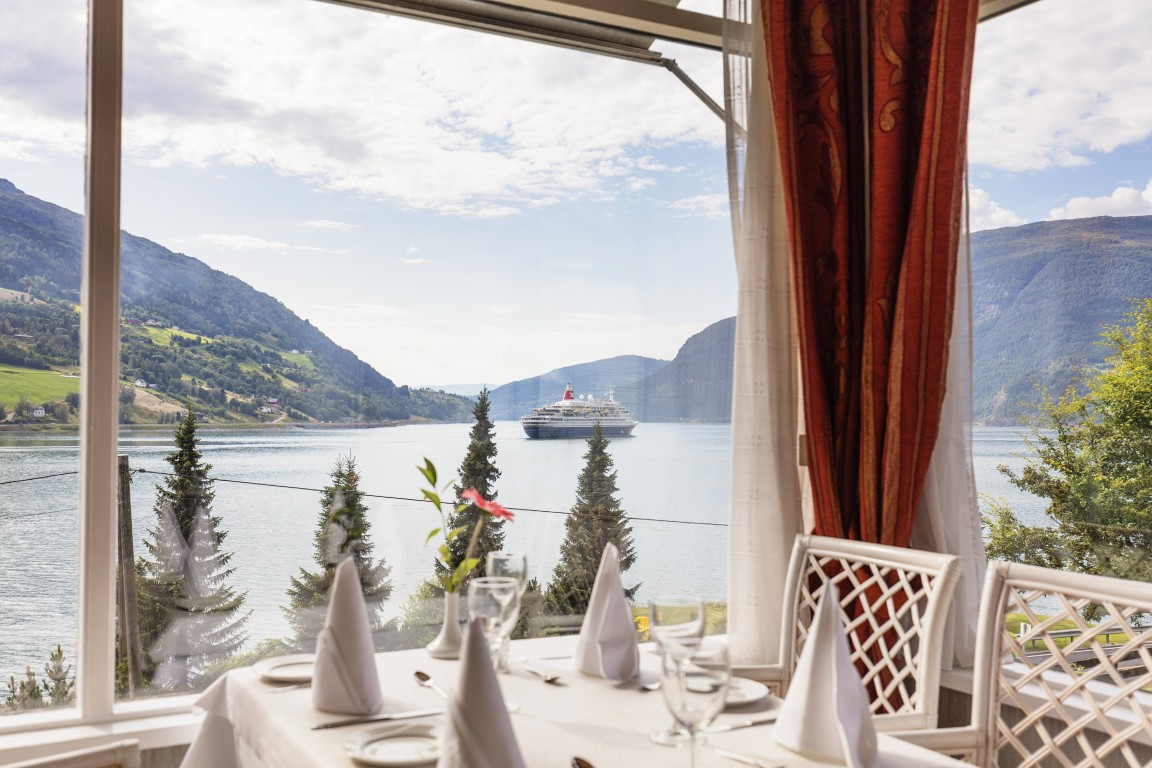 Et dekket bord i restauranten på Olden Fjordhotel. Store vinduer gir en fantastisk utsikt over den blå fjorden. Hvite stoler og røde gardiner gir en klassisk matopplevelse.