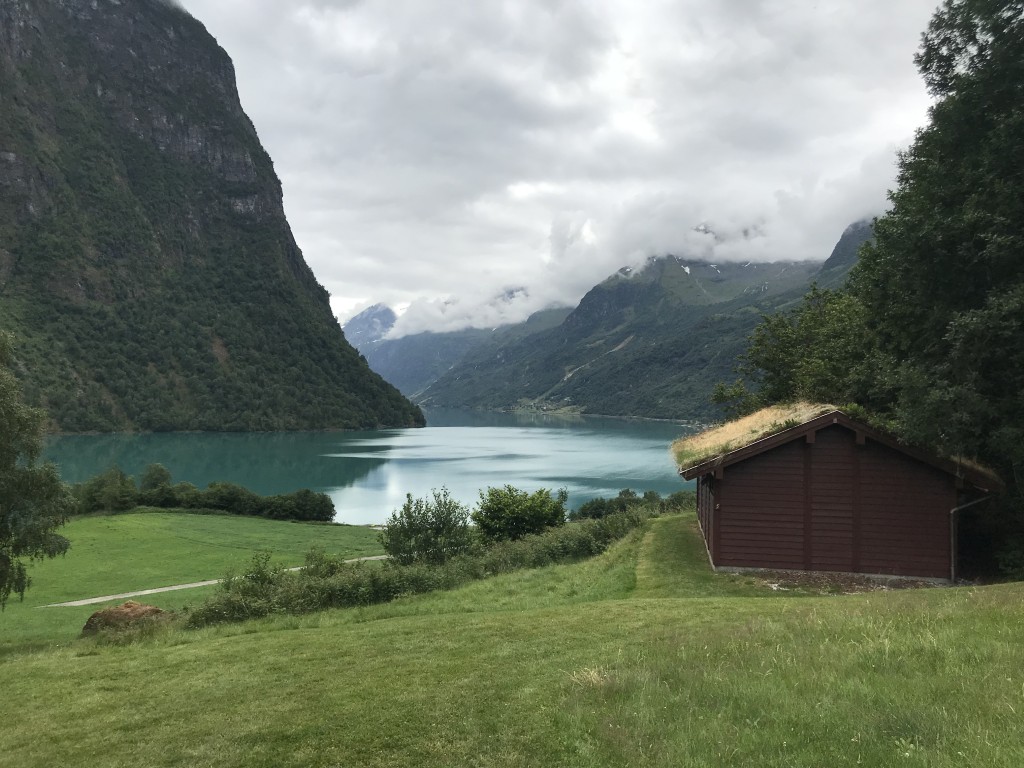 En grønn plen som møter det blå vannet i innsjøen. Flåten feriehus sees til høyre. Gress som vokser fra taket på hytta.