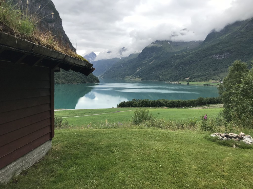 En grønn plen med et hus nær det klare vannet ved Flåten i Olden. Fjellene forsvinner inn i skyene.