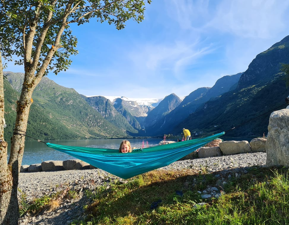 En person ligger i en knallblå hengekøye på Gryta Camping i Olden. Solen skinner med blå himmel bak de høye fjellene.