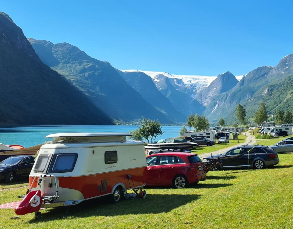 En skitten hvit og rød campingvogn parkert på Gryta Camping i Olden midt på sommeren. Gresset er grønt, fjorden er turkis og himmelblå. Snødekte fjell i bakgrunnen.