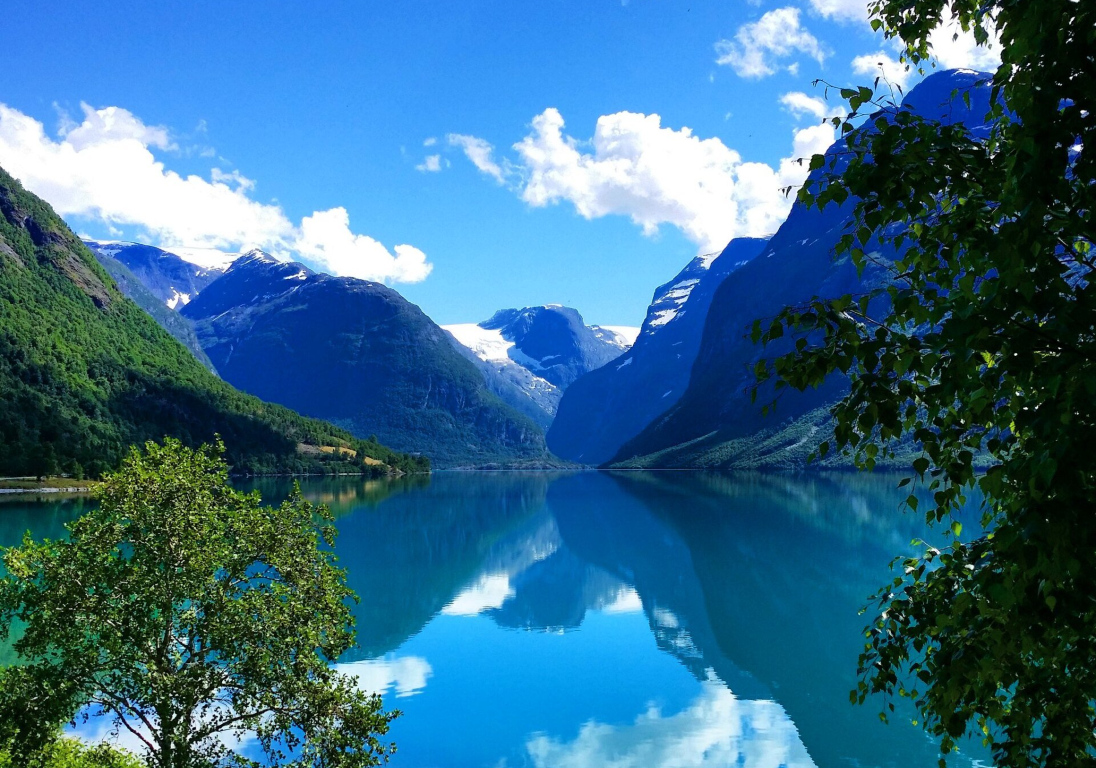Krystallklar innsjø reflekterer de omkringliggende fjellene og den blå himmelen på Gryta Camping. Innrammet av grønnbladede trær.