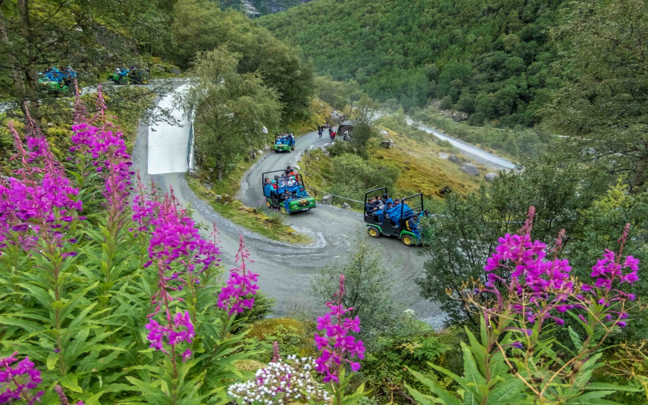 Grønn trollbil foran Briksdalen-breen. To personer står mellom bilen og to høye trær