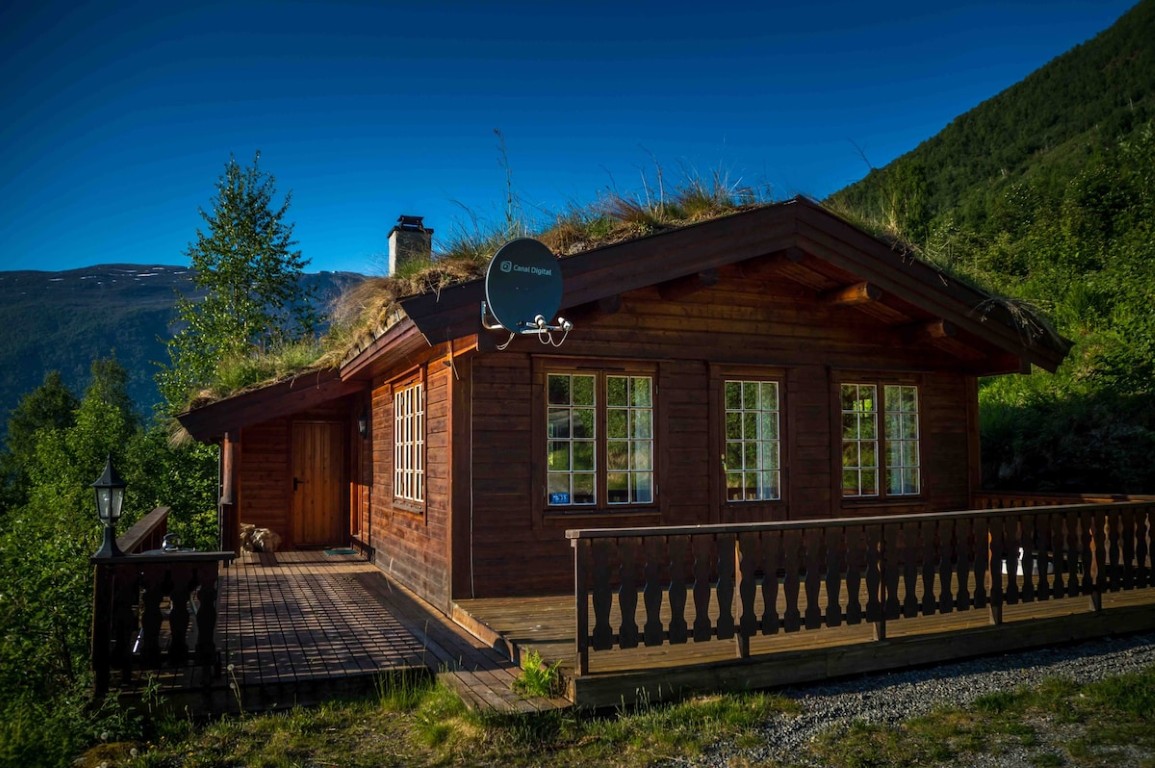 Utvendig av en tradisjonell norsk hytte med gresstak og treterrasse omgitt av sommergrønt og fjell i Olden, Norge