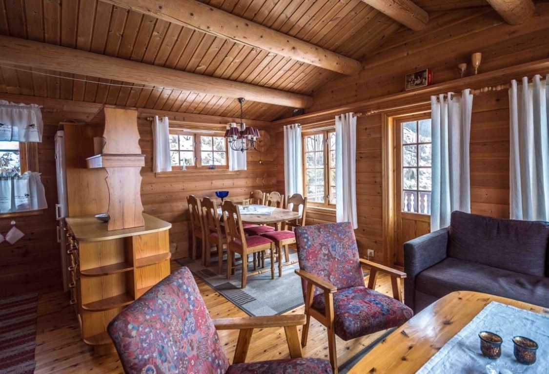 Rustikk åpen stue og spisestue i en norsk hytteutleie nær Olden, med synlige tømmerbjelker, tremøbler og vinduer med fjordutsikt