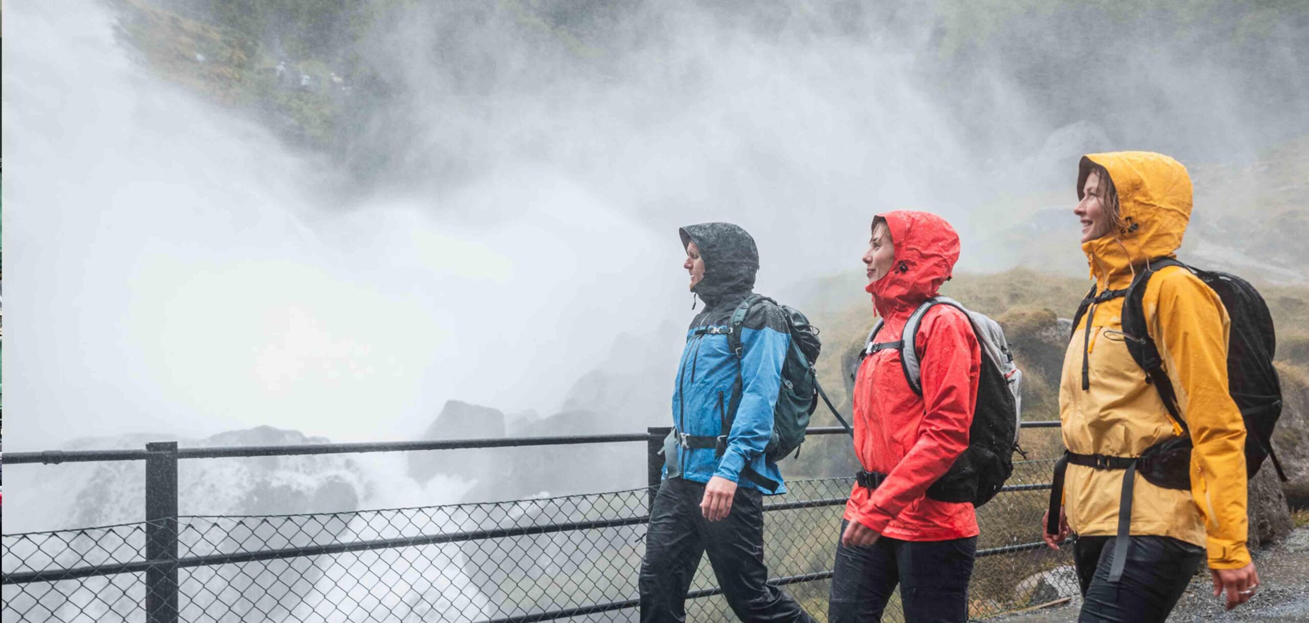 Tre turgåere i Skogstad vanntette jakker går forbi en kraftig foss nær Briksdalsbreen i Olden, Norge